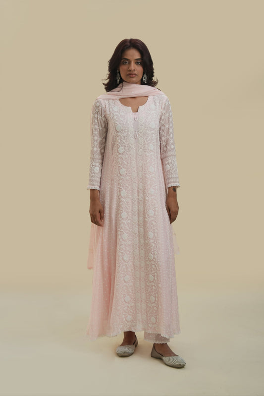 A woman wearing a blush pink Anarkali set with chikankari and mukaish work, paired with a dupatta, standing against a neutral background.