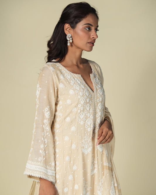 A woman wearing a beige straight tissue suit set with chikankari and mukaish work.