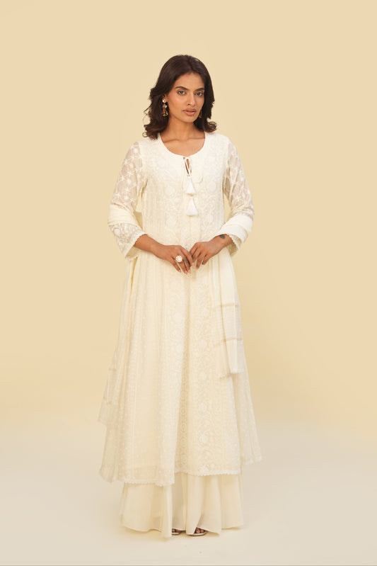 A woman wearing a white Mul Anarkali set with pearl embellishments and tassels on the neckline, standing against a beige background.