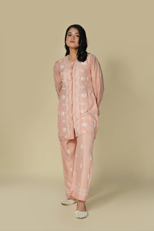 A woman wearing a pink chikankari tissue co-ord set with floral patterns, consisting of a top and pants, standing against a neutral background.