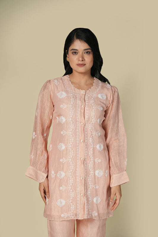 A woman wearing a pink chikankari tissue co-ord set with floral patterns, consisting of a top and pants, standing against a neutral background.