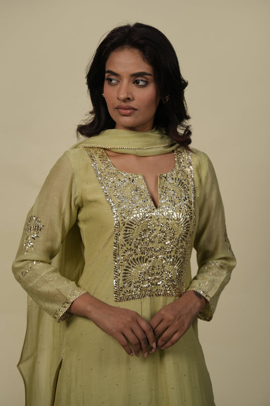 A woman wearing a sage green mukaish silk chanderi suit set with intricate embroidery on the chest area.