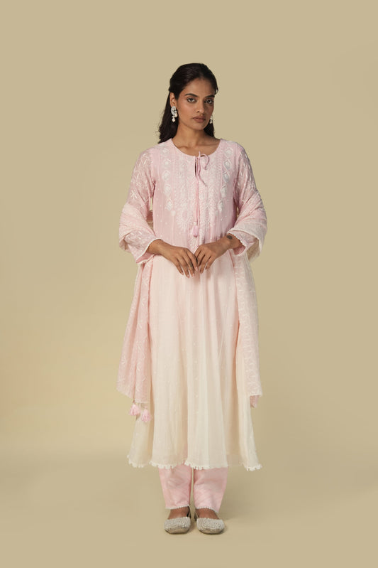 A woman standing against a beige background, wearing a pink anarkali set with chikankari and mukaish detailing.