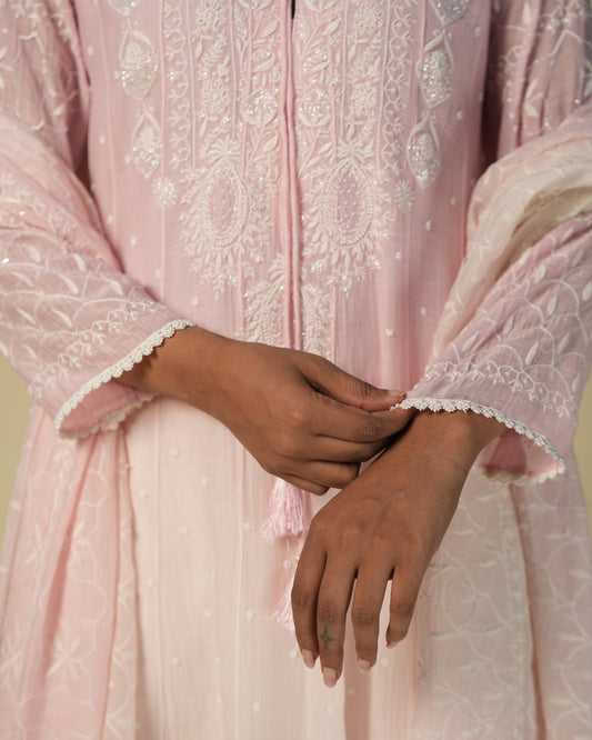 A woman standing against a beige background, wearing a pink anarkali set with chikankari and mukaish detailing.