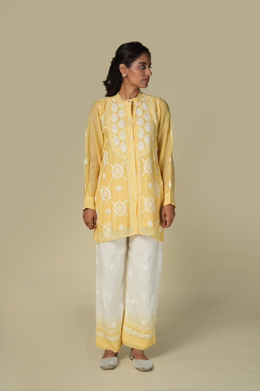 A woman standing in a studio setting, wearing a yellow and white co-ord set with intricate chikankari work on the top, paired with white trousers.