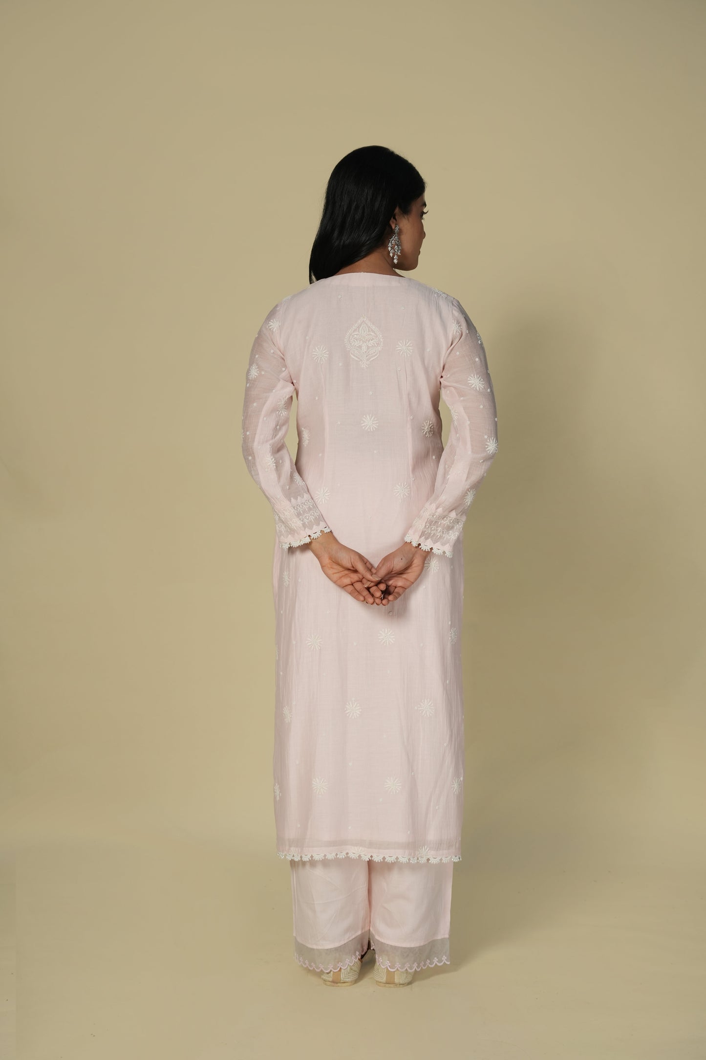 A back view of a woman wearing a baby pink chikankari mul chanderi suit set with delicate embroidery, consisting of a kurta and pants.