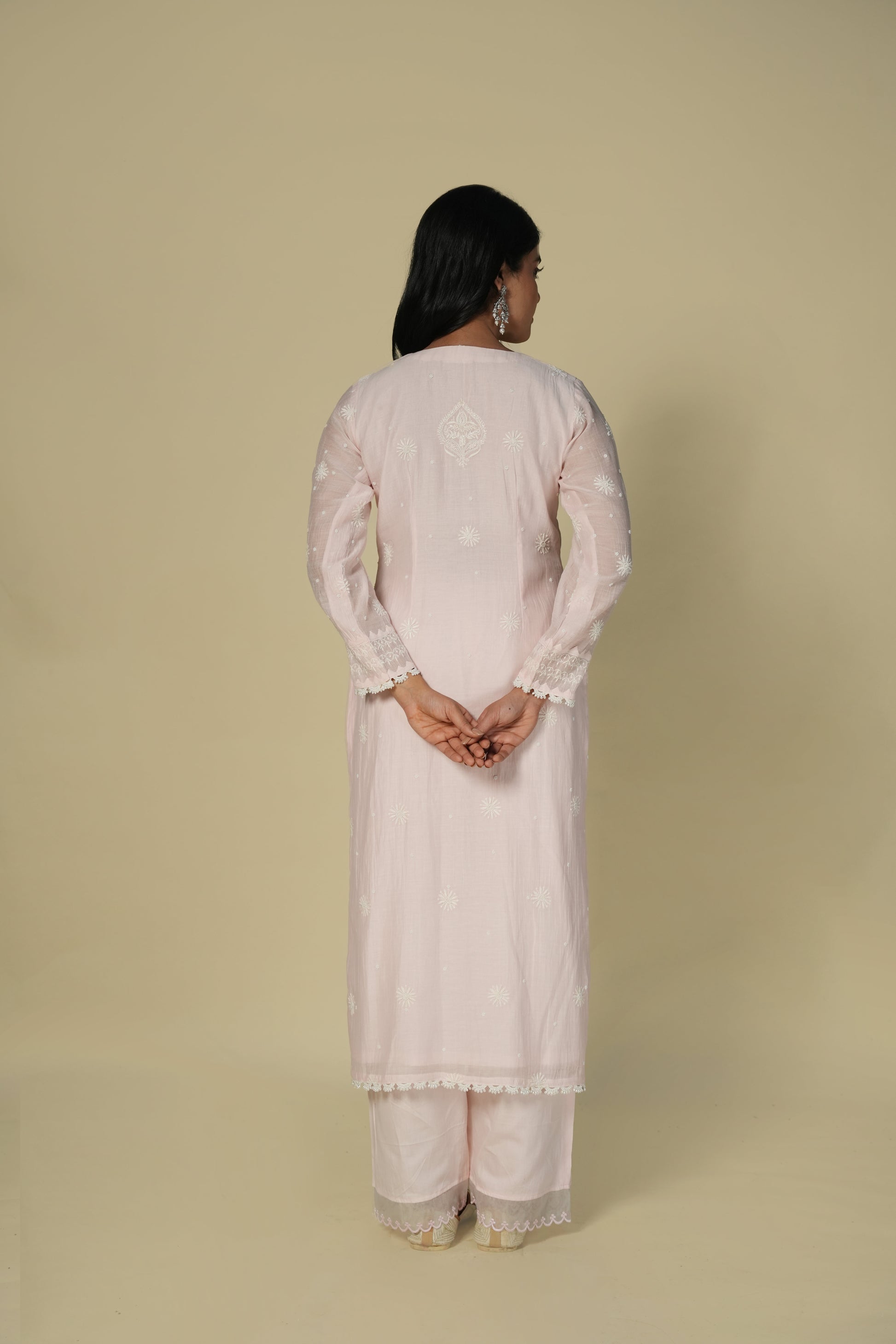 A back view of a woman wearing a baby pink chikankari mul chanderi suit set with delicate embroidery, consisting of a kurta and pants.